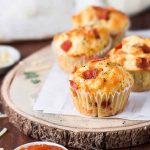 Baked pizza muffins topped with melted cheese, bacon bits, and herbs, served on a wooden board.
