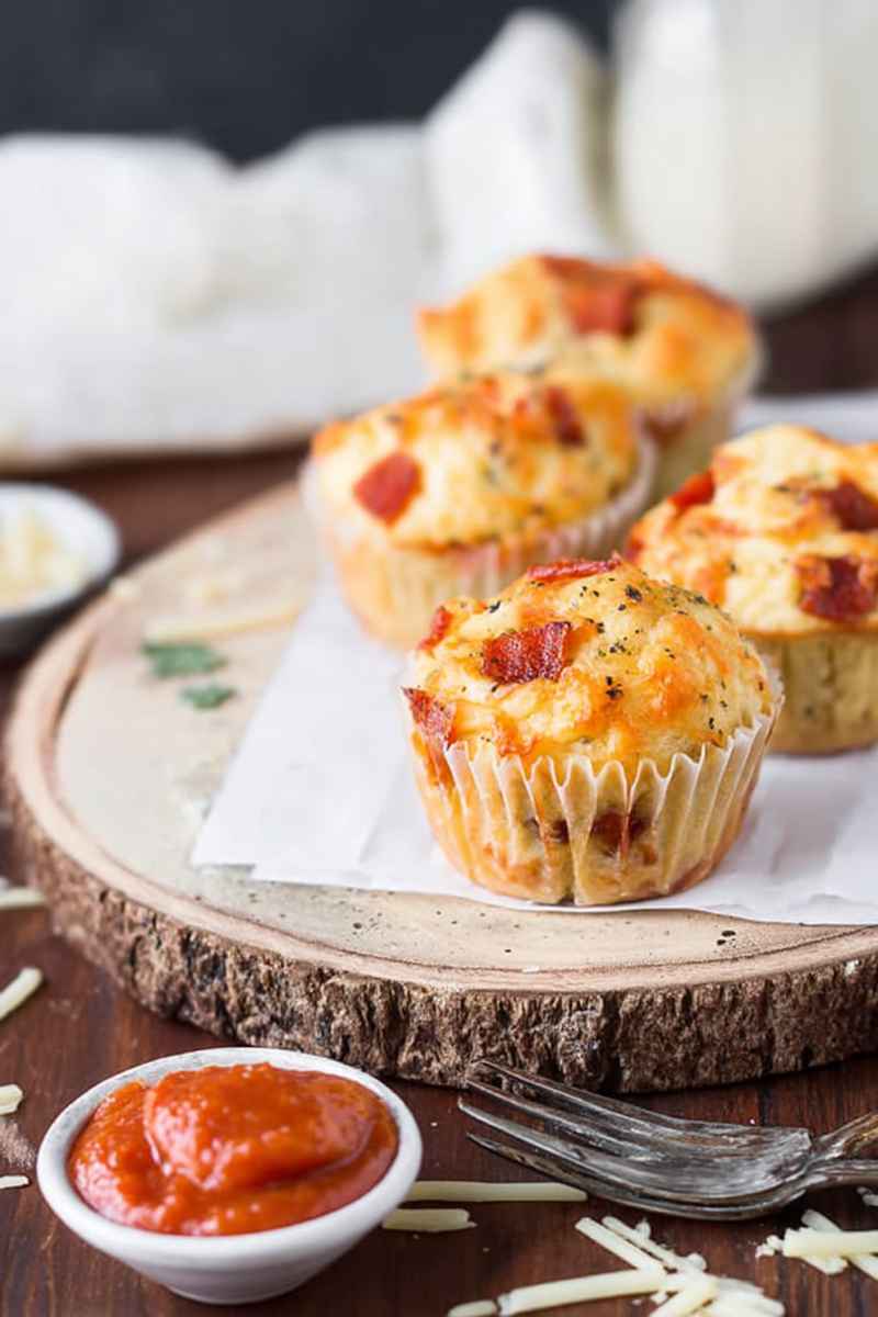 Baked pizza muffins topped with melted cheese, bacon bits, and herbs, served on a wooden board.