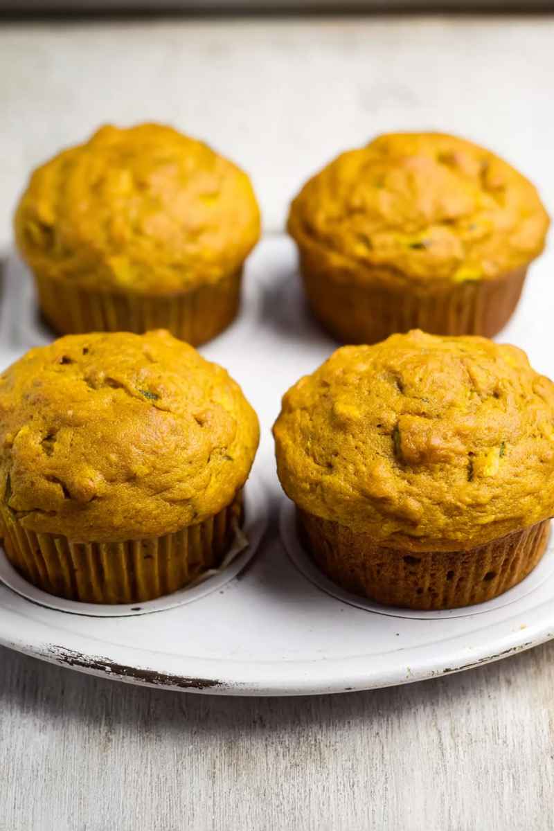 Four golden pumpkin zucchini muffins arranged neatly on a white plate, emphasizing their homemade appeal.
