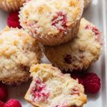 Close-up of a sliced raspberry muffin revealing its moist interior filled with juicy berries, alongside whole raspberries.