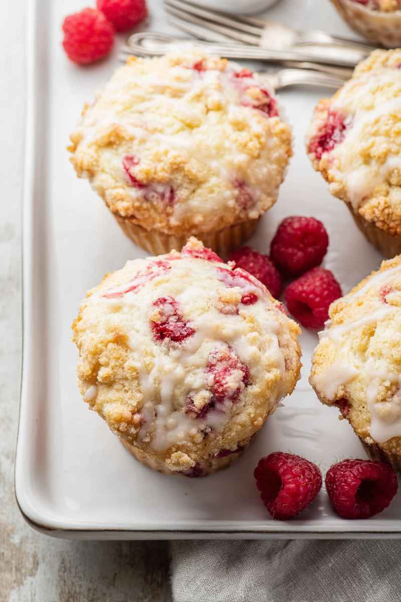 Soft and tender raspberry muffins topped with crumbly streusel and a drizzle of vanilla glaze, surrounded by fresh raspberries.