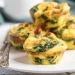 A plate of golden egg muffins topped with melted cheese and flecks of spinach and bacon, presented on a rustic wooden surface.