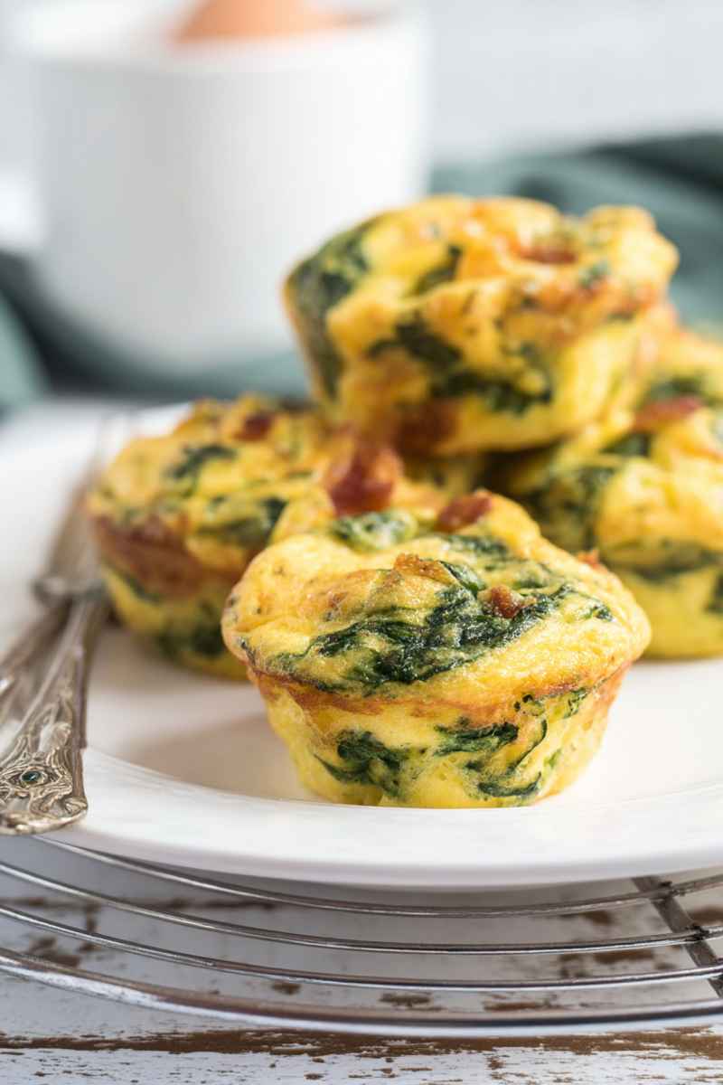 A plate of golden egg muffins topped with melted cheese and flecks of spinach and bacon, presented on a rustic wooden surface.