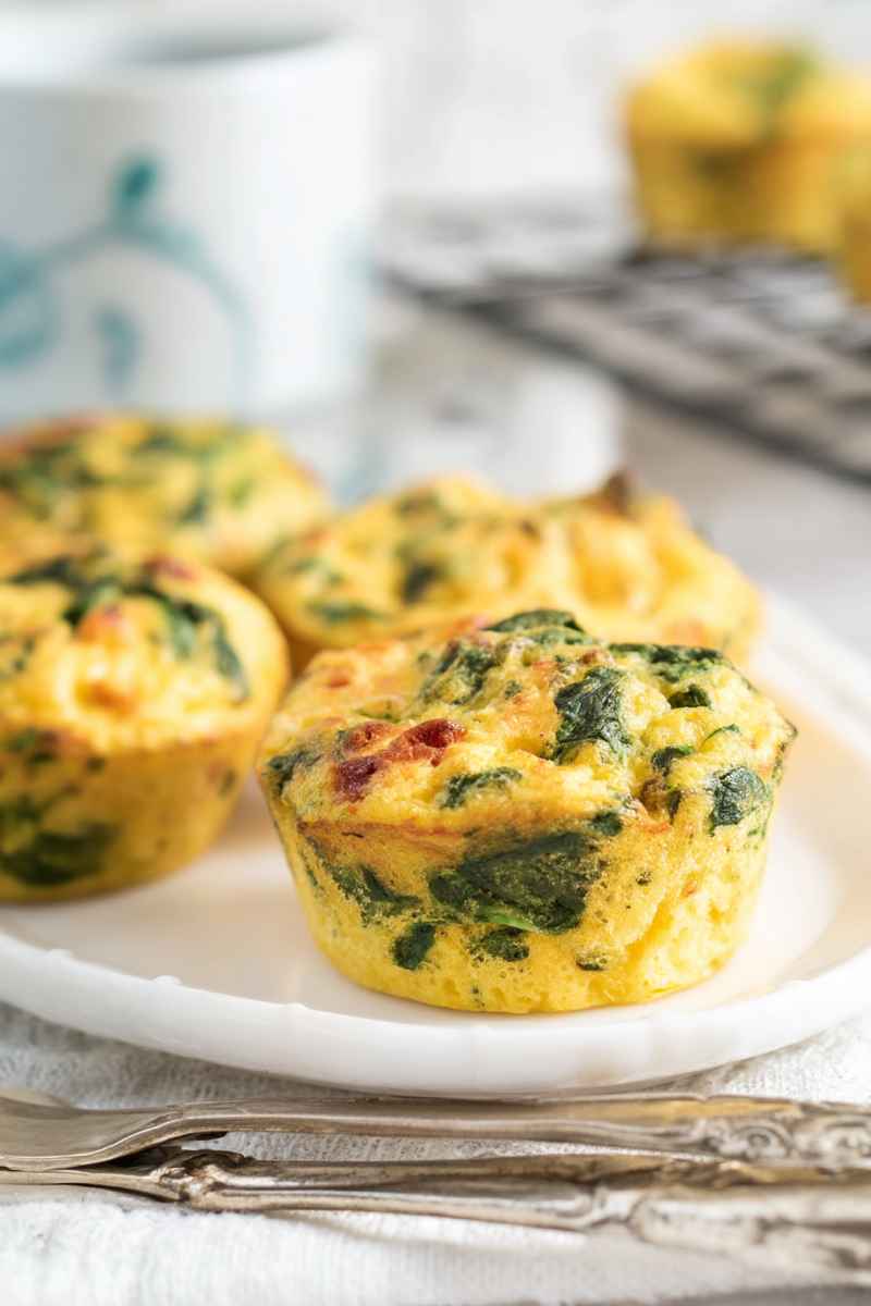 A plate of freshly baked egg muffins with visible layers of spinach and bacon, placed on a white plate with a rustic background.