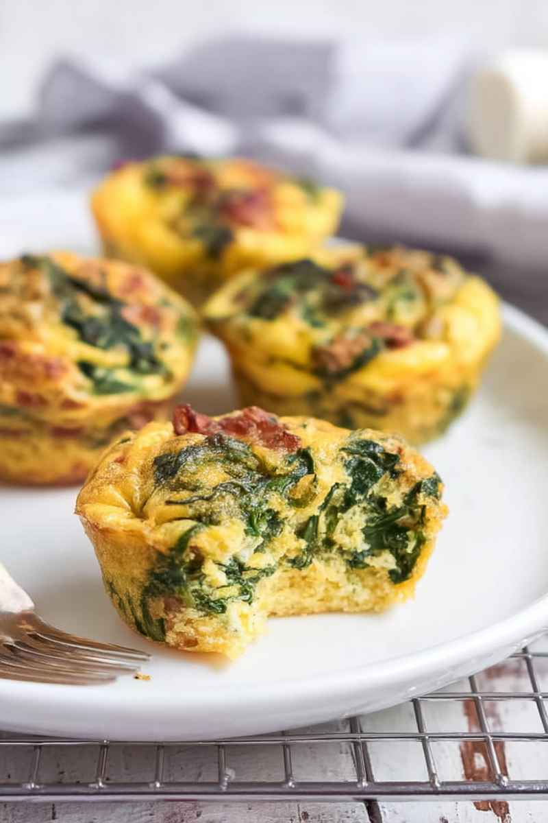 A plate of egg muffins with one cut open, revealing a moist interior filled with spinach and bacon, set against a soft, neutral backdrop.