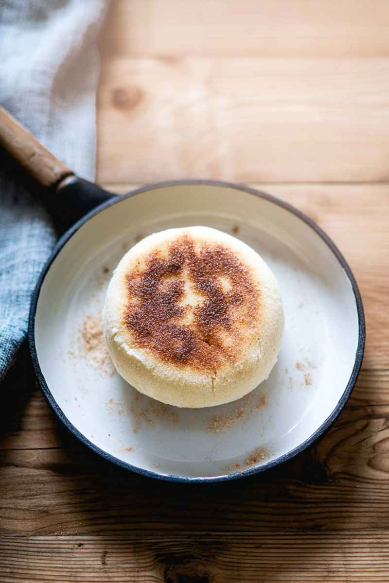 A single sourdough English muffin on a plate, showcasing its golden-brown surface and inviting texture, set against a warm wooden background.