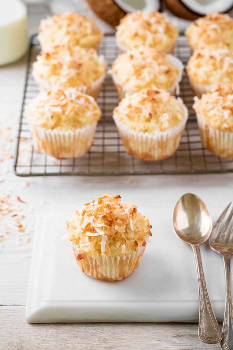 A single toasted coconut muffin on a white plate, with scattered coconut flakes around it, accompanied by vintage silverware.