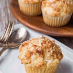 A close-up of a golden toasted coconut muffin on a white plate, with more muffins in the background on a wooden serving board.