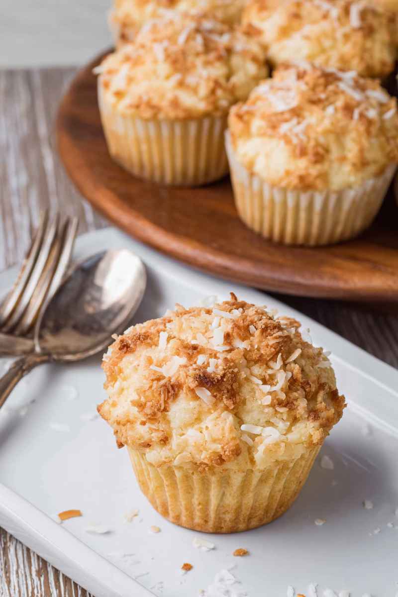 A close-up of a golden toasted coconut muffin on a white plate, with more muffins in the background on a wooden serving board.