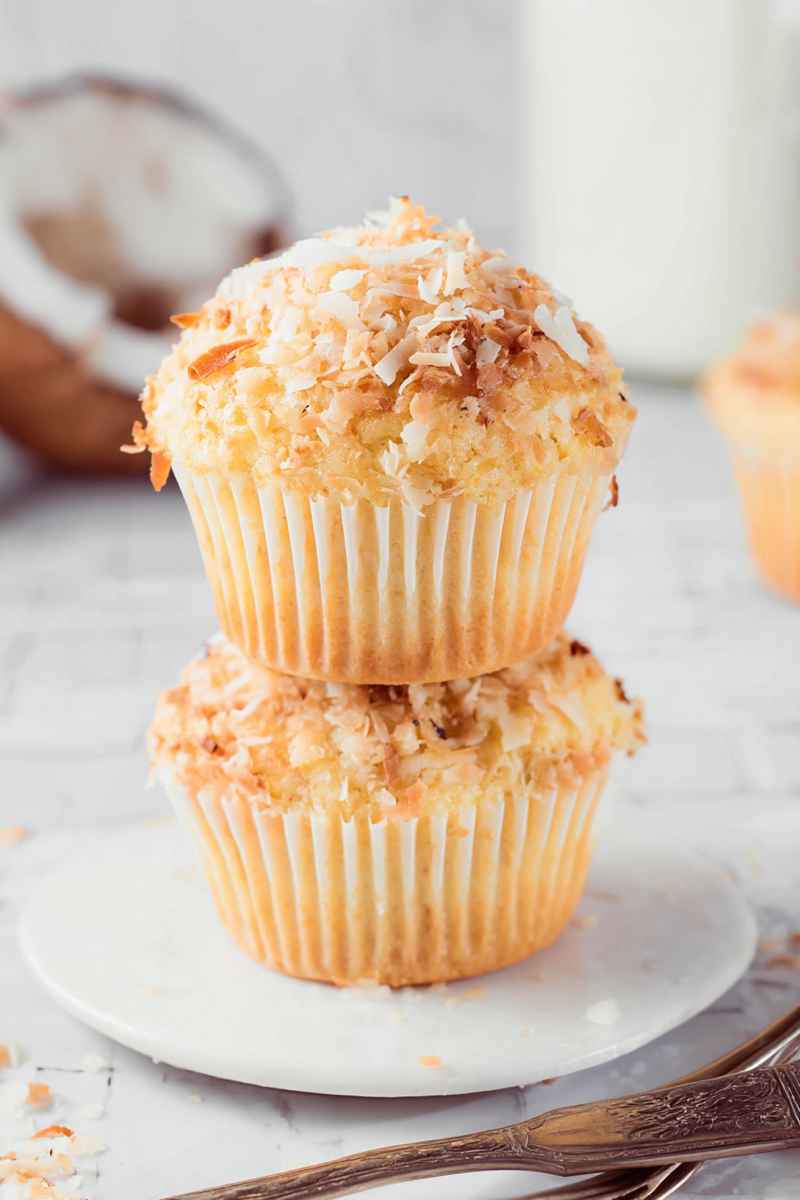 Two golden toasted coconut muffins stacked on a white plate, with scattered coconut flakes and vintage silverware nearby.
