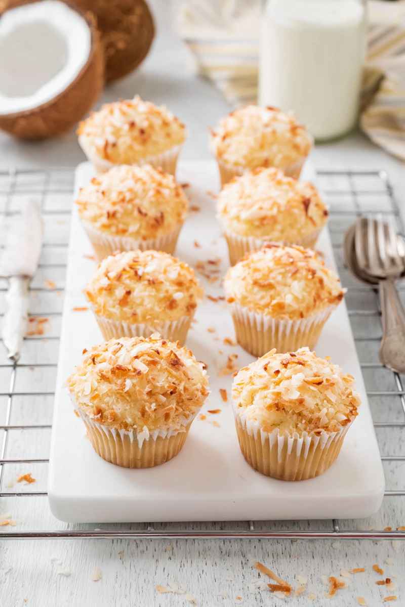 Six freshly baked toasted coconut muffins arranged on a white marble tray, surrounded by a coconut half and a glass of milk.